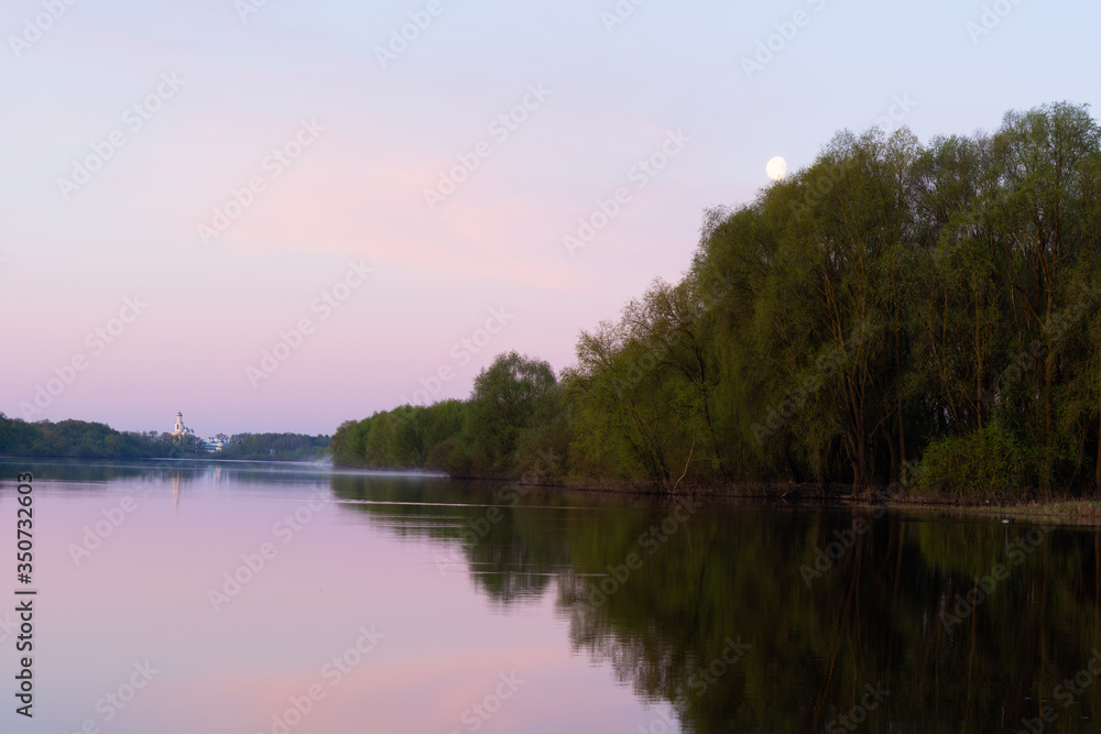 Summer landscape. Willows reflected in the water of the river and the Orthodox monastery in the distance on the horizon and the moon in the sky