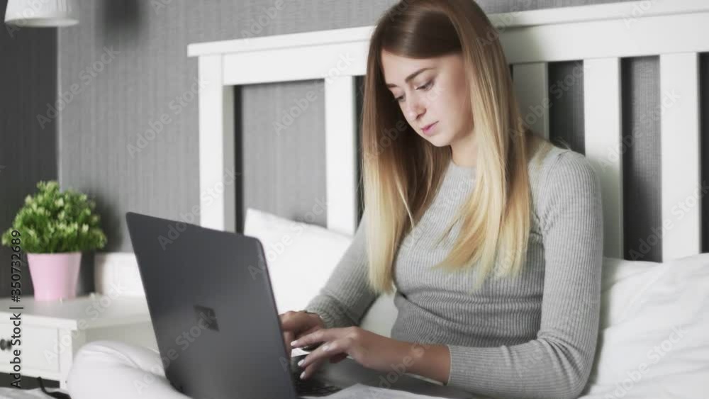 Serious woman sits on bed, uses laptop and works with documents Serious woman sits on bed, uses laptop and works with documents