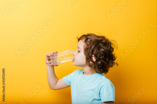 Cute 5 years old curly kid boy drinking clean water from glass at yellow background. clean drinking water concept