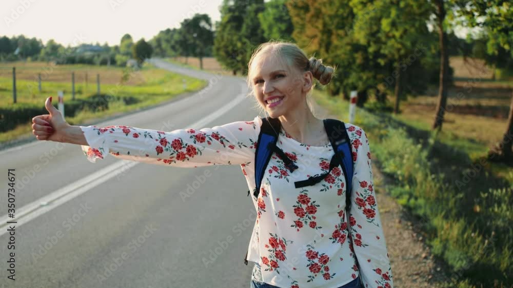 Traveler woman hitchhiking on a sunny road and walking. Young happy backpacker woman looking for a ride to start a journey on a sunlit country road. Traveler woman hitchhiking on a sunny road and walking. Young happy backpacker woman looking for a ride to start a journey on a sunlit country road.