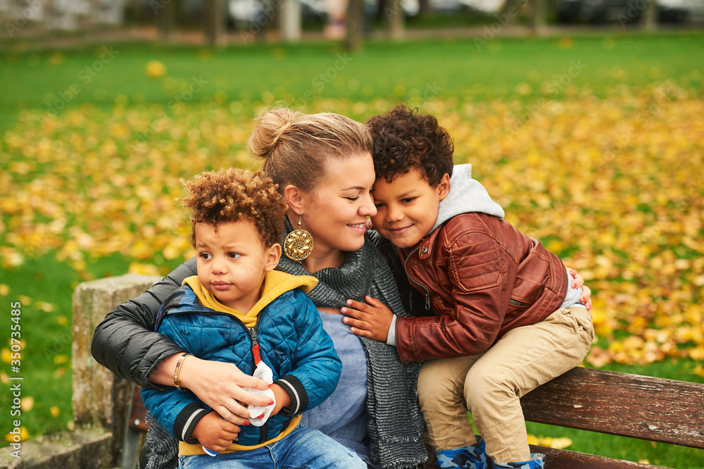 Fototapeta premium Outdoor portrait of happy young mother with two lovely sons, family enjoying nice autumn day in public park, mixed race family