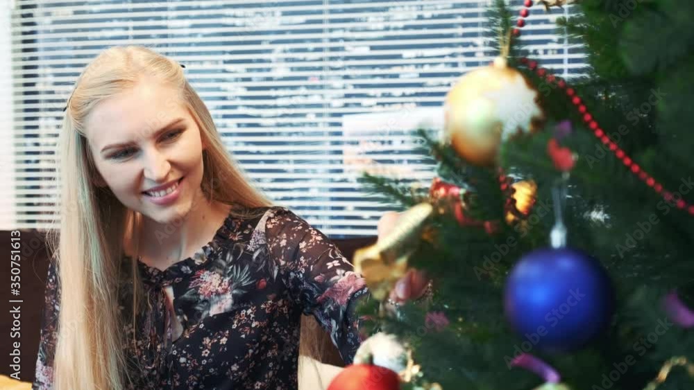 Close up of smiling girl hanging balls on Christmas tree with skyscrapers on the background. The woman smiles and decorates the fir tree with colorful toys. Close up of smiling girl hanging balls on Christmas tree with skyscrapers on the background. The woman smiles and decorates the fir tree with colorful toys.