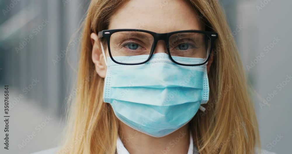 Close up of face of young Caucasian beautiful businesswoman in glasses and medical mask looking straight to camera with happy look. Portrait of woman at street. Coronavirus concept. Close up of face of young Caucasian beautiful businesswoman in glasses and medical mask looking straight to camera with happy look. Portrait of woman at street. Coronavirus concept.