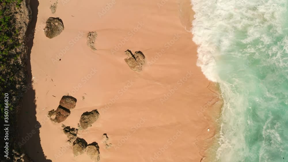 Top down aerial view from above of giant ocean waves crashing and foaming on empty sand tropical beach with big rock stones. Bird's eye aerial shot of golden beach meeting deep blue ocean water. Top down aerial view from above of giant ocean waves crashing and foaming on empty sand tropical beach with big rock stones. Bird's eye aerial shot of golden beach meeting deep blue ocean water.