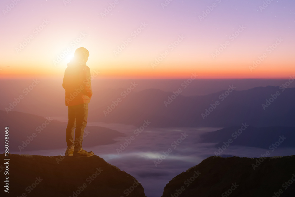 Silhouette of young traveler wearing red sweater standing alone on top of the mountain enjoy looks at the beautiful view sunrise and foggy landscape. The concept of tourism travel and male loneliness.