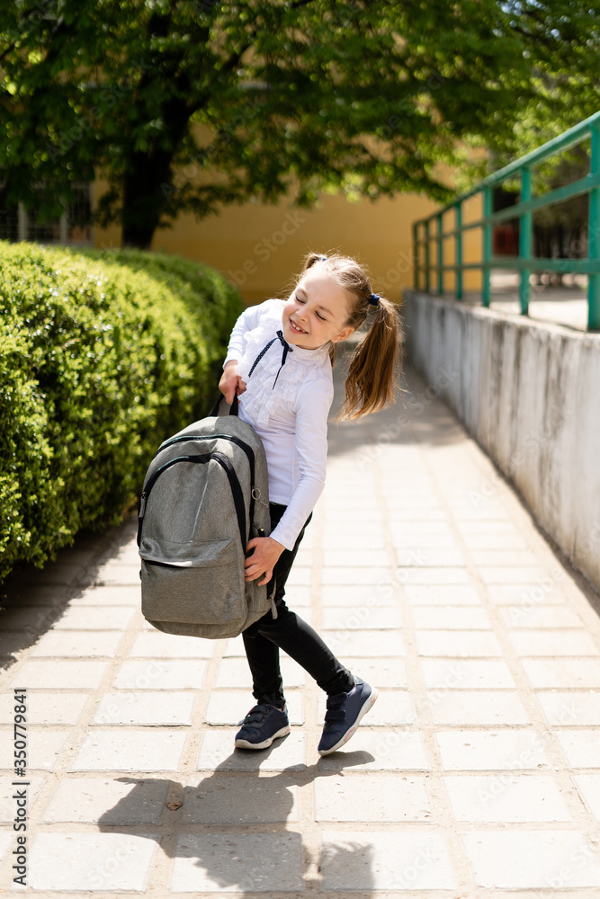 Beautiful young girl holding a heavy backpack. The child returns to school. There are books and notebooks in the bag. The student smiles and goes to study. Stock Photo Adobe Stock