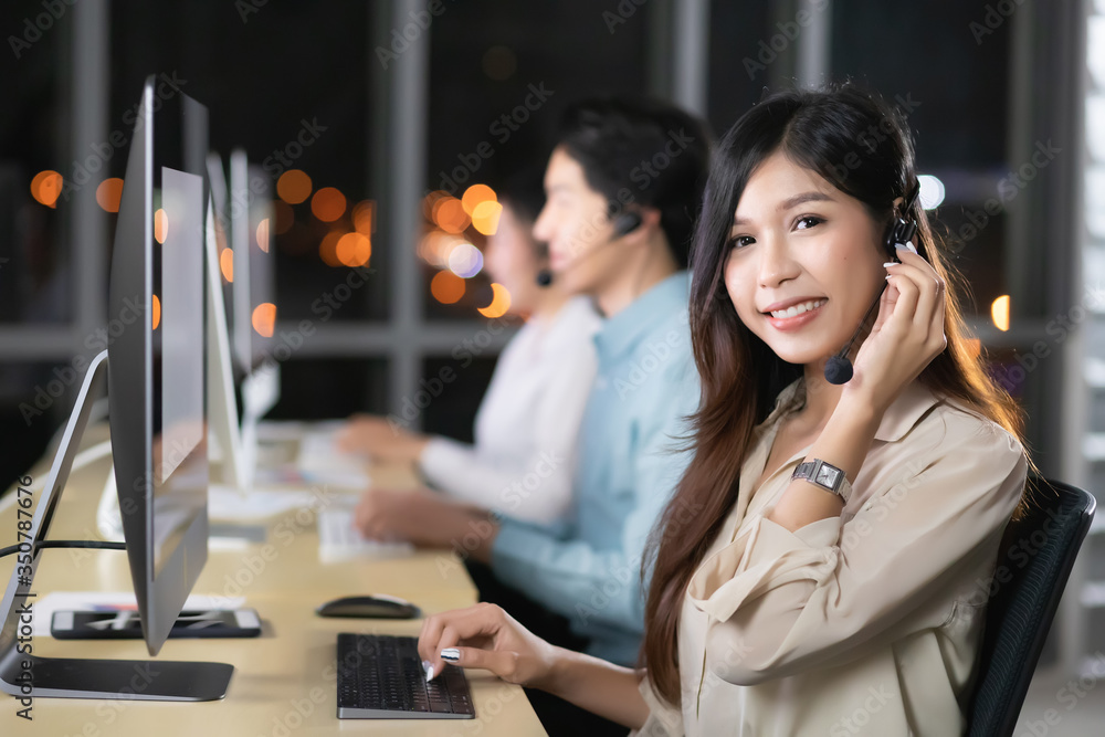 A beautiful Asian woman, Thai, is wearing her work uniform and headphones sitting in front of the computer. She is smiling and grasping the headphones in the after sales and customer service center A beautiful Asian woman, Thai, is wearing her work uniform and headphones sitting in front of the computer. She is smiling and grasping the headphones in the after sales and customer service center