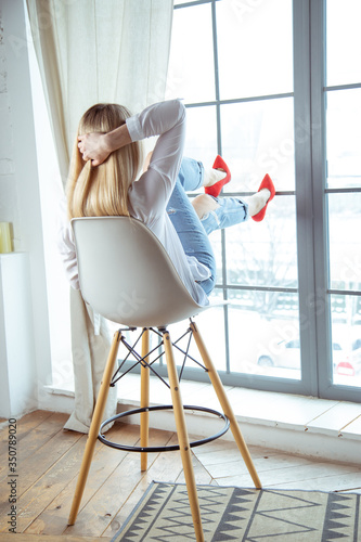 the girl is sitting beautifully on a high chair near a large window, red shoes, high heels. I put my foot on the glass