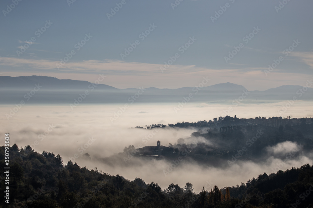Surreal view of of a little town in Umbria (Italy) almost completely hidden by fog with trees silhouettes in the foreground