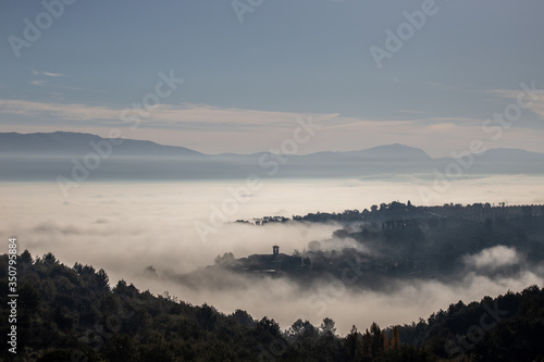 Surreal view of of a little town in Umbria (Italy) almost completely hidden by fog with trees silhouettes in the foreground © Massimo