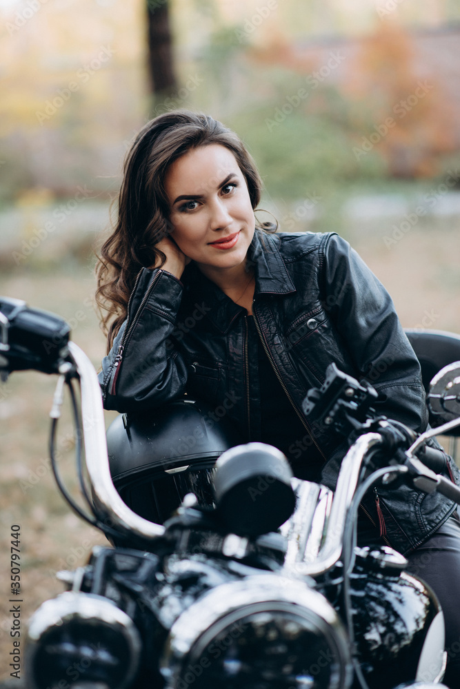 Young beautiful biker woman in a black leather jacket on a motorcycle with a helmet in his hands. The concept of speed and freedom. Soft selective focus. Young beautiful biker woman in a black leather jacket on a motorcycle with a helmet in his hands. The concept of speed and freedom. Soft selective focus.
