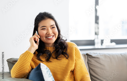 people, technology and communication concept - happy smiling asian young woman in yellow sweater sitting on sofa and calling on smartphone at home