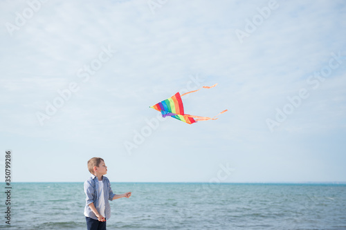 funny little caucasian boy with flying multicolored kite on blue sky and sea background with copy space