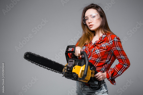 Girl with a chainsaw on a gray background. Girl in a red shirt and construction safety glasses