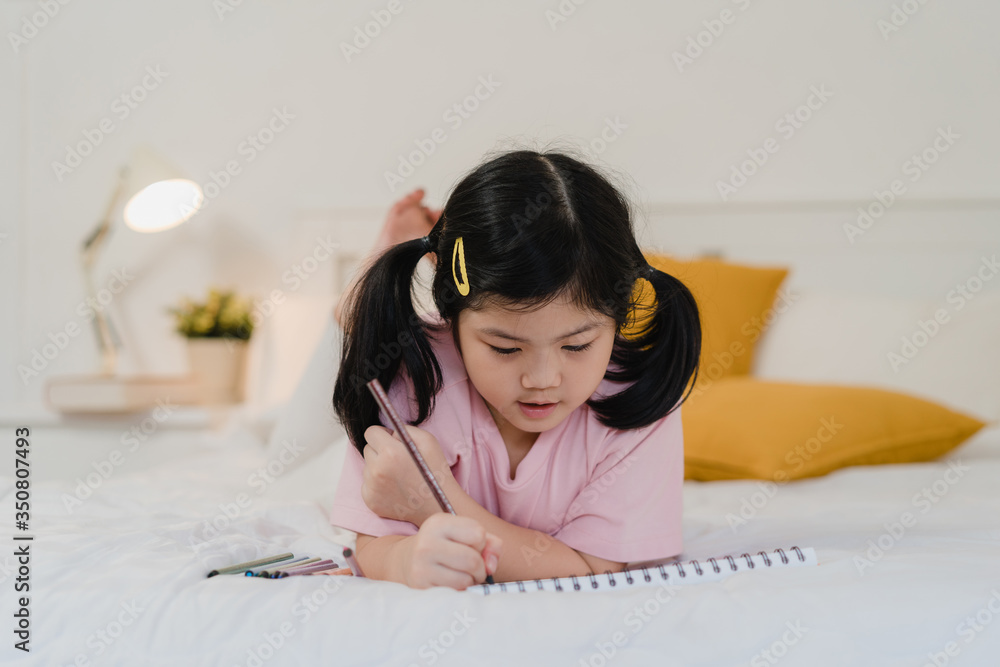Naklejka premium Young Asian girl drawing at home. Asia japanese woman child kid relax rest fun happy draw cartoon in sketchbook before sleep lying on bed, feel comfort and calm in bedroom at night concept.