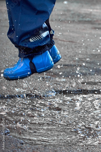 Happy child jumping in puddle in waterproof coat. A boy have fun in the rain weather in a bright raincoat. Legs of child with rubber boots.