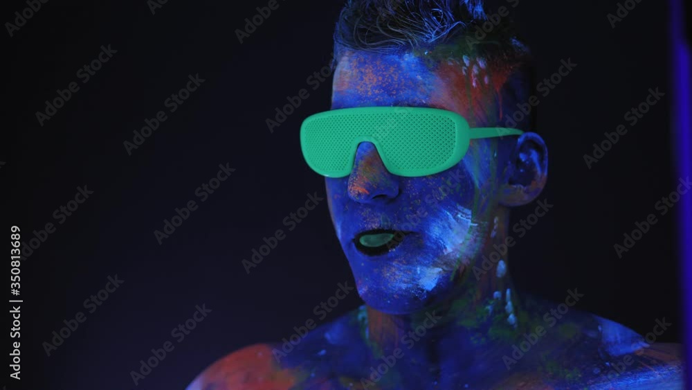 A man with UV drawings on his face and green neon glasses inflates a bubble of bubblegum in the dark under fluorescent lighting. Bright fluorescent body art glows in darkness. UV paint. A man with UV drawings on his face and green neon glasses inflates a bubble of bubblegum in the dark under fluorescent lighting. Bright fluorescent body art glows in darkness. UV paint.