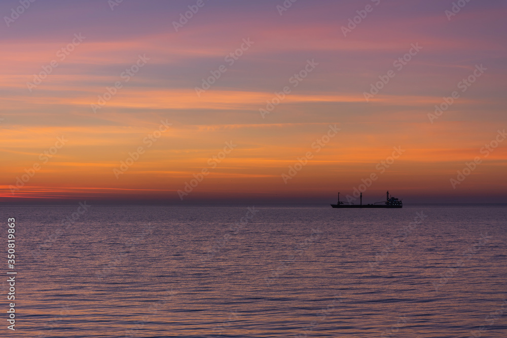 Bright twilight sunset sky over the sea. Wonderful sky after sunset in orange shades. The natural background. Purple hue of the sea and sky. Abstract lines of fire clouds. A ship on the horizon.