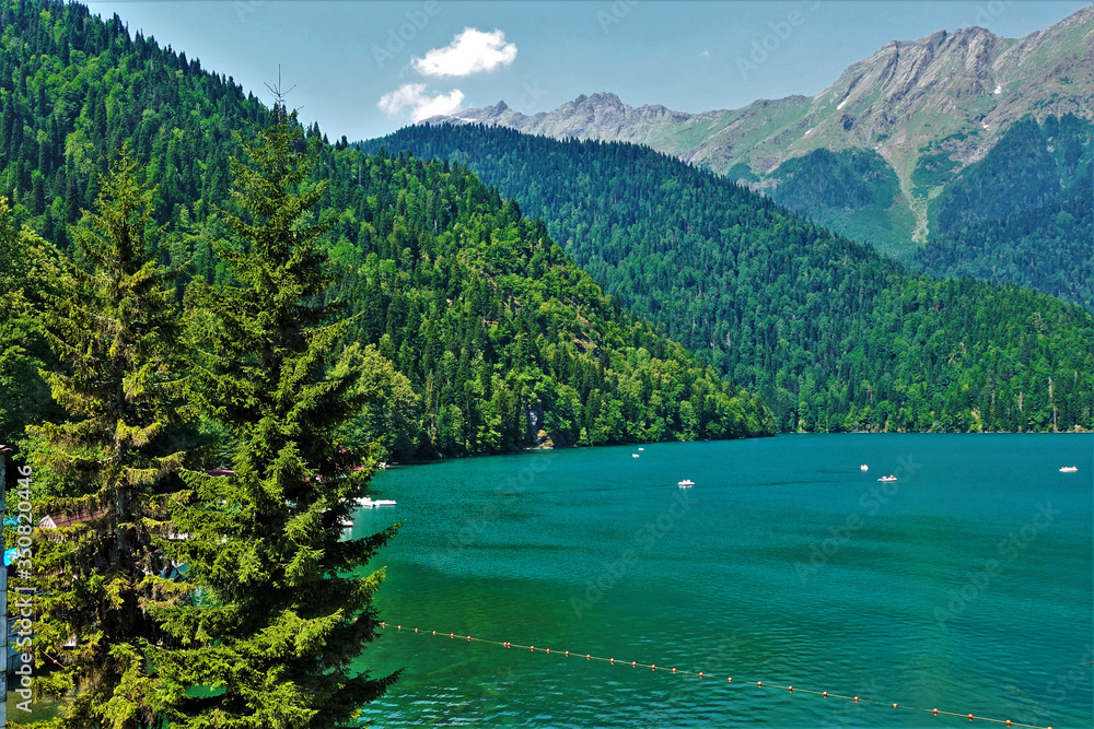 Unique beautiful alpine lake Ritsa. Abkhazia. Boats float on calm aquamarine water. The surrounding mountains are covered with emerald forest. In the sky above the picturesque cliffs light clouds.
