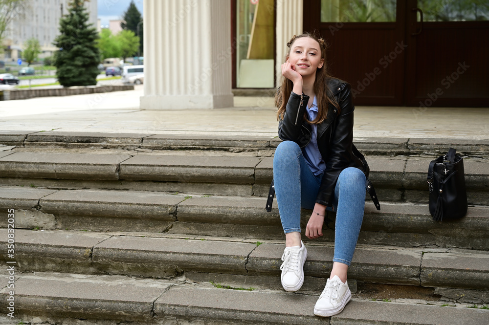 Model posing sitting in spring park outdoors in the city on the steps. Photo of a young pretty girl with a smile in a black jacket and jeans.