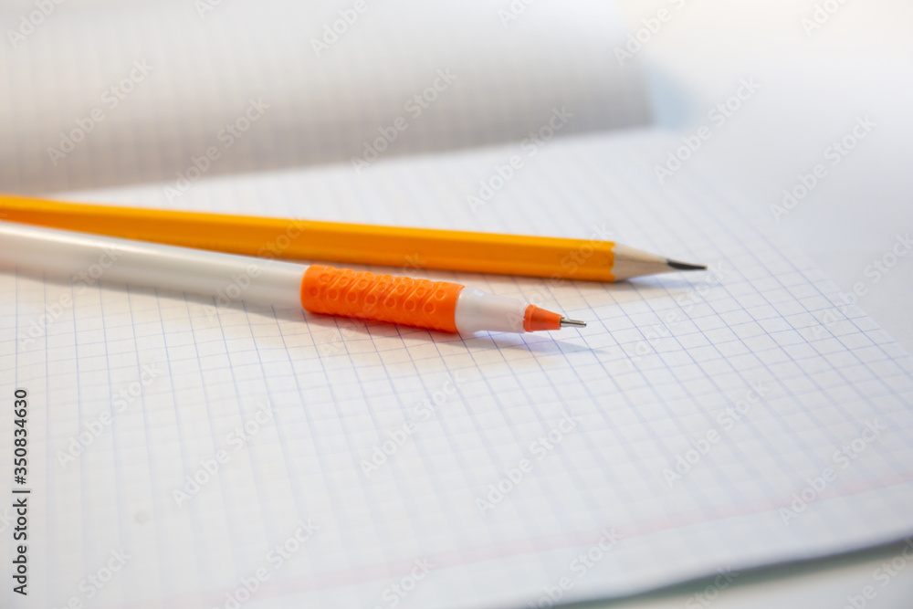 School tools. Orange pen and yellow pencil lie on on a notebook in a cage. Office and school tools. School tools. Orange pen and yellow pencil lie on on a notebook in a cage. Office and school tools.