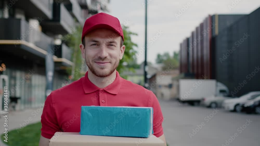 Portrait handsome smiling young courier postman holding cardboard boxes parcels standing in business street district residential area outdoors. Home delivery concept. Portrait handsome smiling young courier postman holding cardboard boxes parcels standing in business street district residential area outdoors. Home delivery concept.