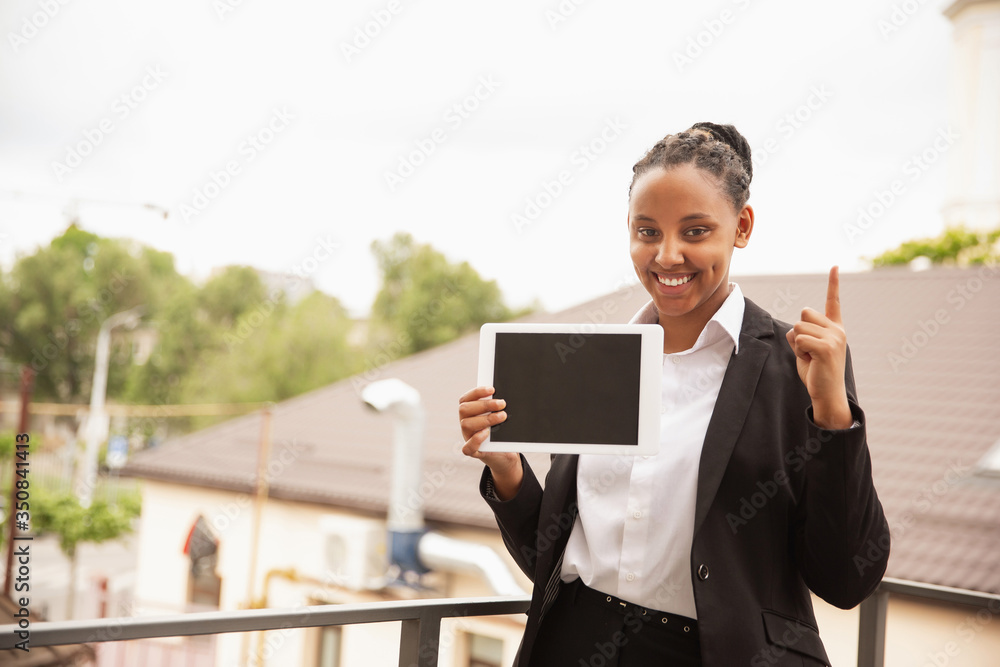 Pointing up with tablet. African-american businesswoman in office attire smiling, looks confident and happy. Finance, business, equality and human rights concept. Beautiful young model, successful. Pointing up with tablet. African-american businesswoman in office attire smiling, looks confident and happy. Finance, business, equality and human rights concept. Beautiful young model, successful.