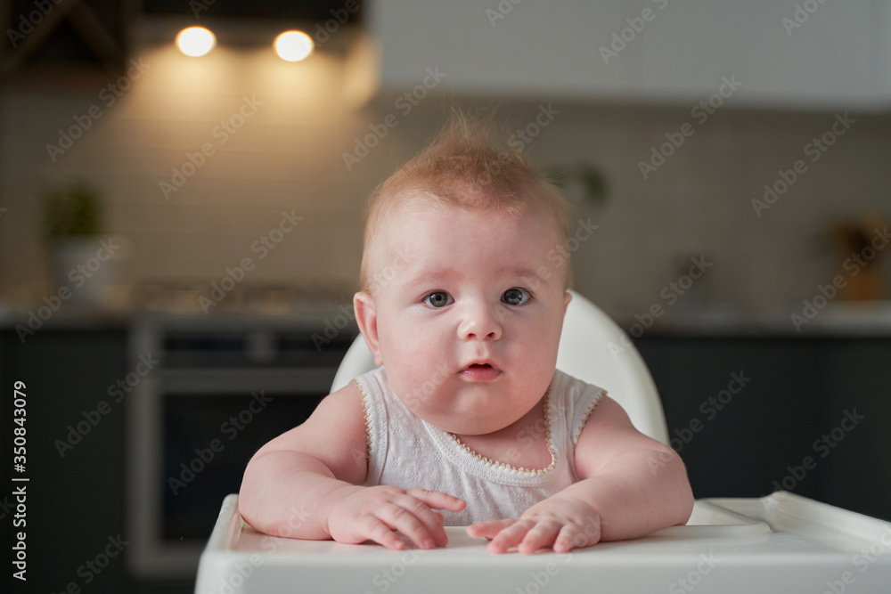 cute blond child sits in a children's chair at the kitchen and eat a juicy peach. Little girl with a piercing look