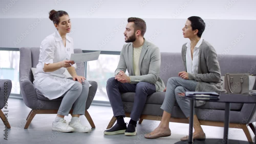 Medium shot of male patient with beard showing jaw x-ray to female dentist in white coat while having consultation in reception area of dental office. Supportive wife sitting on couch beside him Medium shot of male patient with beard showing jaw x-ray to female dentist in white coat while having consultation in reception area of dental office. Supportive wife sitting on couch beside him