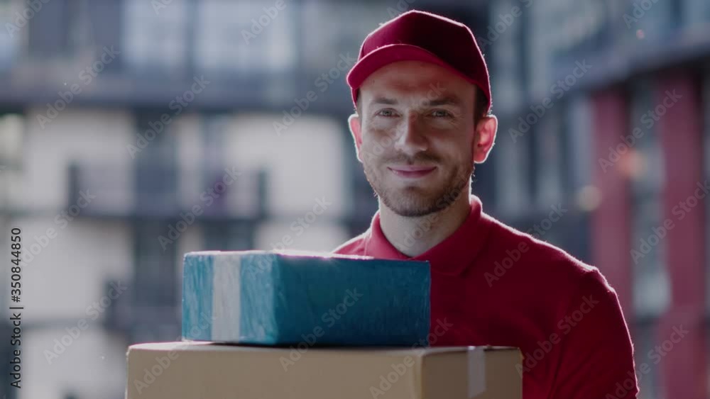 Outdoor portrait of handsome courier young postman smiling cheerful holding customer parcel cardboard boxes waiting on street in contemporary residential district cityscape. Outdoor portrait of handsome courier young postman smiling cheerful holding customer parcel cardboard boxes waiting on street in contemporary residential district cityscape.