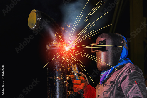A young man welder in brow uniform, welding mask and welders leathers, weld metal with arc welding machine with sparks light on black background.
