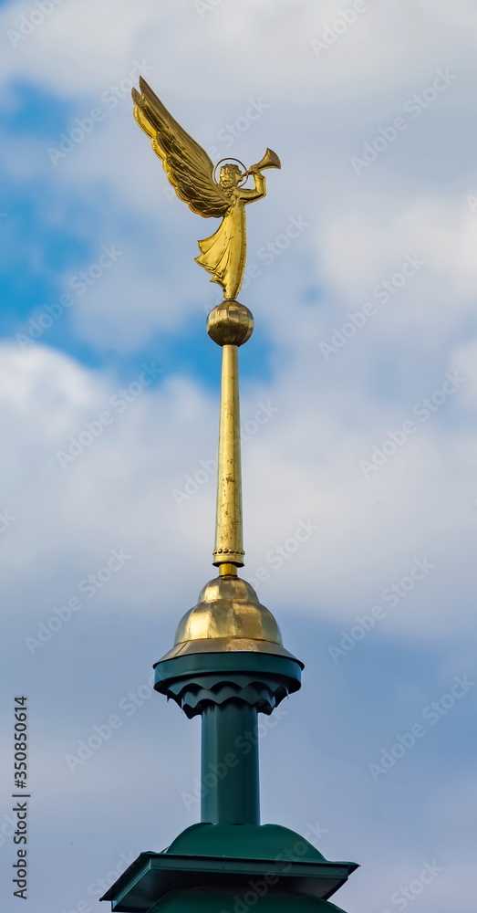 Gilded angel with a trumpet on the spire of the bell tower of the Orthodox Church against the blue sky with clouds closeup
