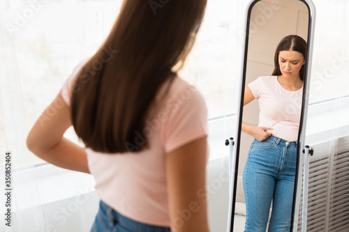 Unhappy Woman Gaining Weight Touching Belly Standing At Mirror Indoors photo