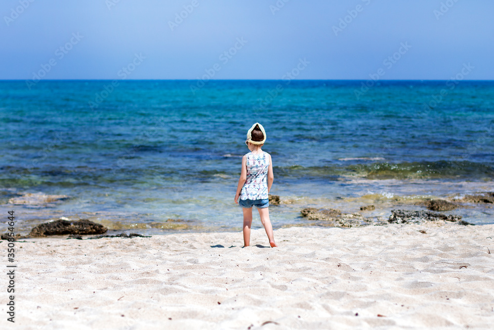 Little girl on an empty white beach looks into the sea during summer vacation