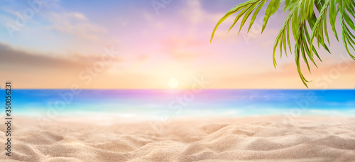 A summer vacation  holiday background of a tropical beach and blue sea at sunset with the sun on the horizon and green palm tree leaves.
