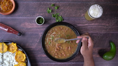Above view of a black man’s hand stirring bean curry in a rustic artisanal bowl on a set table including a rice dish with fried plantain and fresh naranjilla juice