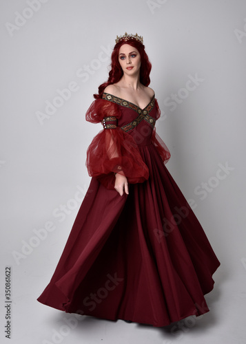 Portrait of a beautiful woman with red hair wearing a flowing Burgundy fantasy gown and golden crown. full length standing pose, isolated against a studio background 