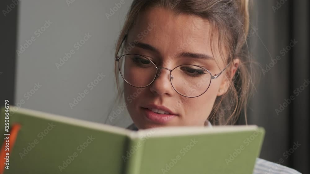 A close-up view of a smiling young woman wearing glasses is making notes in her notebook sitting on the sofa at home  