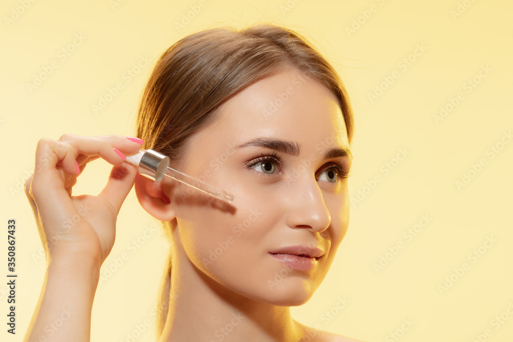 Close up of beautiful young woman with pouring serum, oil on yellow background. Concept of cosmetics, makeup, natural and eco treatment, skin care. Shiny and healthy skin, fashion, healthcare. Close up of beautiful young woman with pouring serum, oil on yellow background. Concept of cosmetics, makeup, natural and eco treatment, skin care. Shiny and healthy skin, fashion, healthcare.