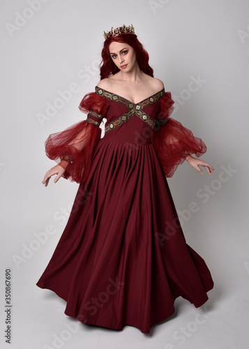 Portrait of a beautiful woman with red hair wearing  a  flowing Burgundy fantasy gown and golden crown.  full length standing pose  isolated against a studio background 