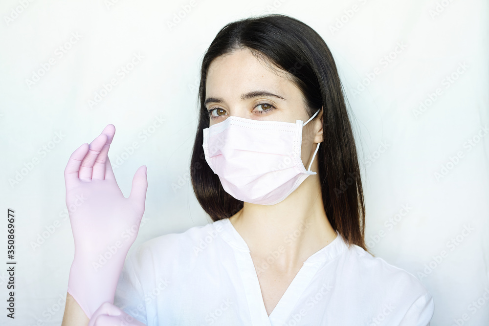 A woman in a pink protective mask adjusts her pink gloves. The girl stands on a light background in white clothes. The concept of suppression of coronavirus