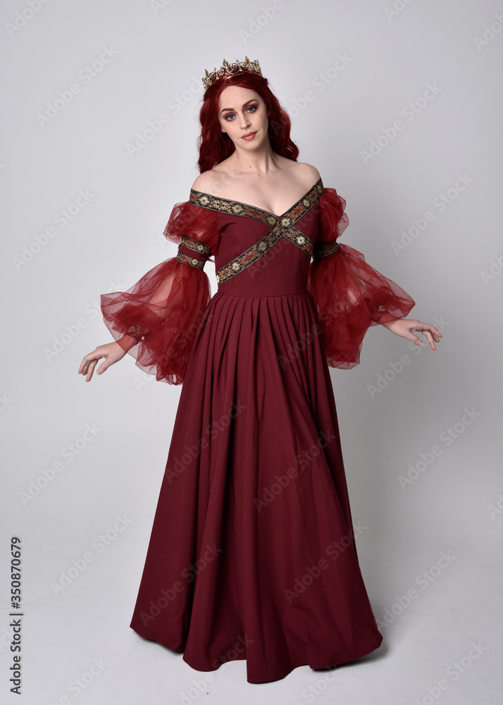Portrait of a beautiful woman with red hair wearing  a  flowing Burgundy fantasy gown and golden crown.  full length standing pose, isolated against a studio background
