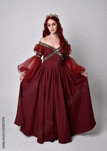 Portrait of a beautiful woman with red hair wearing a flowing Burgundy fantasy gown and golden crown. full length standing pose, isolated against a studio background 