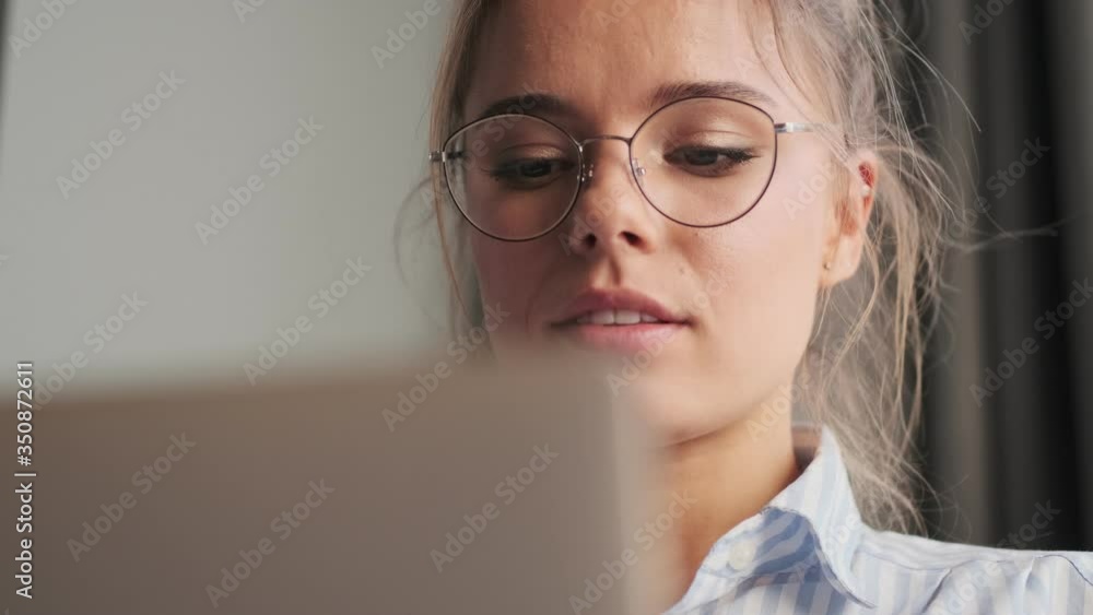 A close-up view of a beautiful young woman wearing glasses is using her laptop while sitting in the living room at home A close-up view of a beautiful young woman wearing glasses is using her laptop while sitting in the living room at home