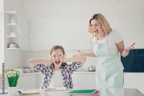 Photo of two people crazy furious mother shout her small daughter sit table cant write essay avoid close cover ears palms yell wear dotted apron in house kitchen indoors