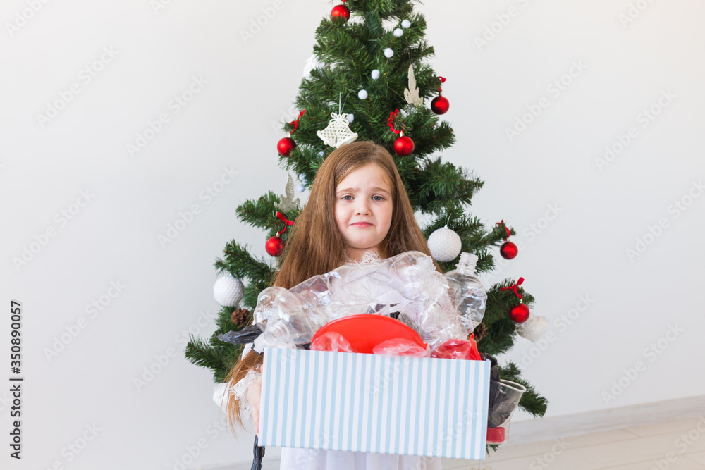 Shocked little child girl looks with opened eyes and worried expression, holding box with various plastic wastes over christmas tree background. Shocked little child girl looks with opened eyes and worried expression, holding box with various plastic wastes over christmas tree background.