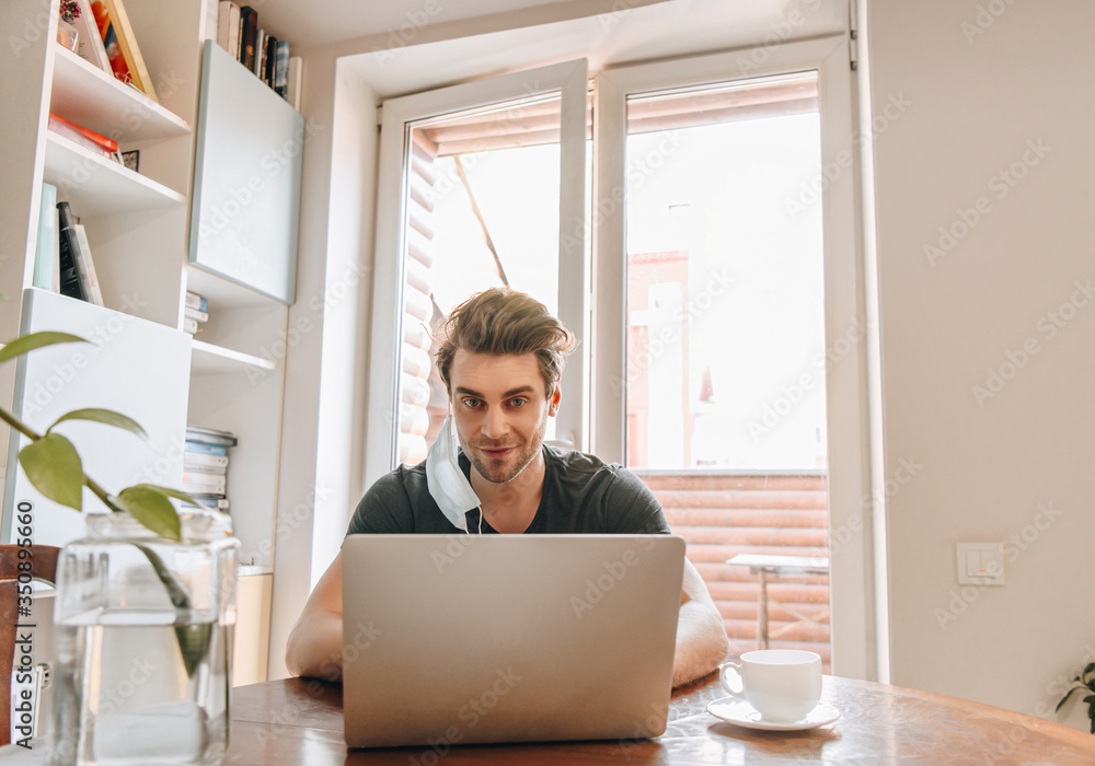 smiling man with medical mask hanging on ear working on laptop near cup of coffee smiling man with medical mask hanging on ear working on laptop near cup of coffee