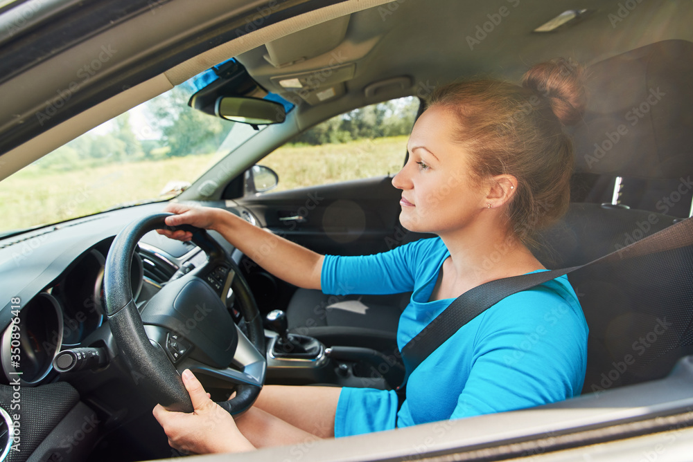 Obraz premium Happy young woman in blue dress driving a car, side view. Beautiful girl at the wheel of car with black interior on the road