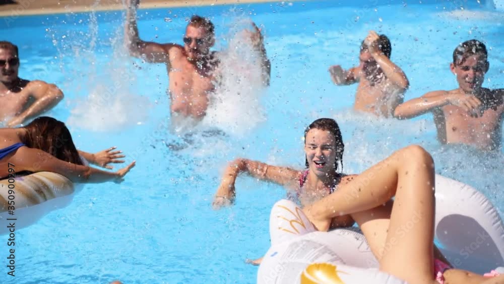 Friends have party in a private villa swimming pool. Happy young people in swimwear splashing water, dancing with floaties and inflatable mattress in luxury resort on sunny day. Tracking shot. Friends have party in a private villa swimming pool. Happy young people in swimwear splashing water, dancing with floaties and inflatable mattress in luxury resort on sunny day. Tracking shot.