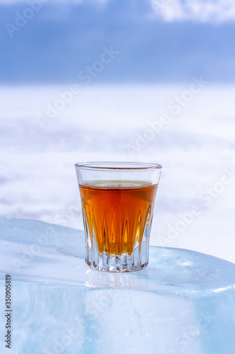 An orange alcoholic drink in a glass stands on an ice piece. Ice melted in the sun. Light blurred background. Copy space. Side view. Vertical.